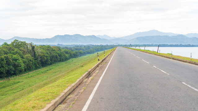 A long empty road runs along the edge of Udawalawe Reservoir, bordered by green grassland and forest with misty hills in the background - Powered by Adobe