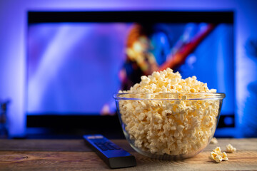 A glass bowl of popcorn and remote control in the background the TV works. Evening cozy watching a movie or TV series at home