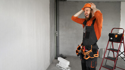 Male bearded builder in hard hat and blue overalls with working tools on belt has problem holding...