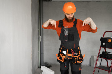 Male bearded builder in hard hat and blue overalls with working tools on belt say look there Pointing Fingers down at copy space. Repair, construction and building