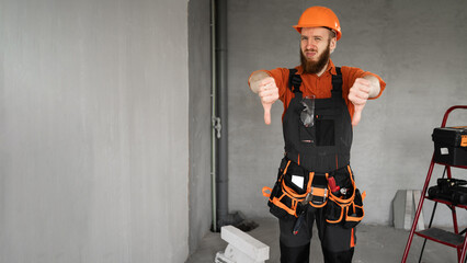 Unhappy Male bearded builder in hard hat and blue overalls with working tools on belt showing thumbs-down dislike gesture. Repair, construction and building