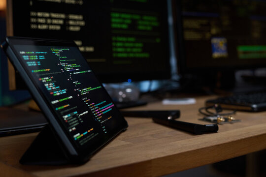 Tablet displaying colorful programming code on wooden desk with stylus and earphones resting nearby, computer monitors with code in background suggesting software development workspace