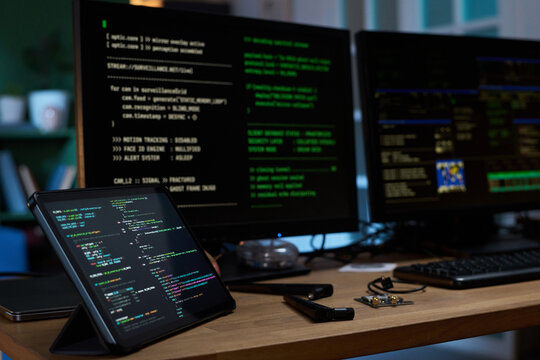 Closeup showing digital tablet and two computer monitors displaying programming code on wooden desk in workspace, electronic components and stylus lying nearby, no people