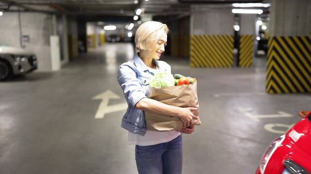 A woman smiles while loading a bag of fresh groceries into the trunk of her car in a parking garage