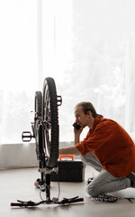 Young long-haired man wearing orange shirt talking on phone with mechanic, asking for help to...
