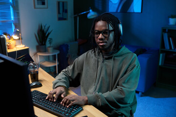 Young Black man wearing headphones sitting at desk using desktop computer typing on keyboard in home office environment with plants and lamp in background