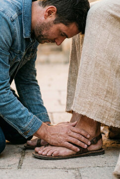 A man in a denim jacket kneeling in humility and resting his forehead against the linen robe of Jesus while touching his feet in a gesture of worship and surrender.