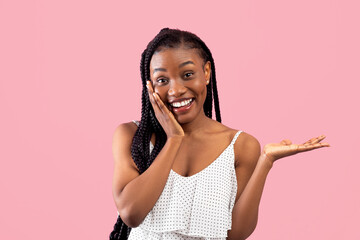 Amazed black lady touching her face and demonstrating something over pink studio background. Charming African American woman advertising your product, promoting sale or discount
