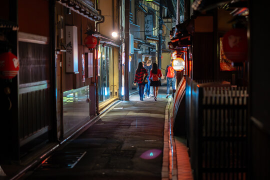 View of warm light spills onto a narrow alleyway as pedestrians stroll past traditional buildings with red lanterns, Kyoto, Kyoto, Japan.