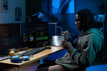 Young Black man sitting at desk working on computer with multiple monitors displaying code, holding bowl and eating with spoon, wearing glasses, focused on screen in office