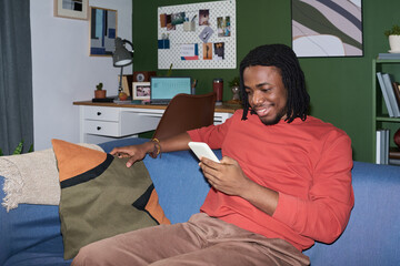 Young Black man sitting on sofa using smartphone, smiling while reading message, relaxed posture...
