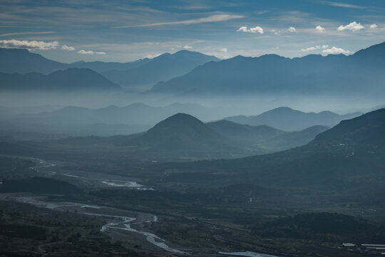 View of layers of hazy blue mountains receding into the distance, with a winding river cutting through the green valley below, Trikala, Thessaly, Greece.