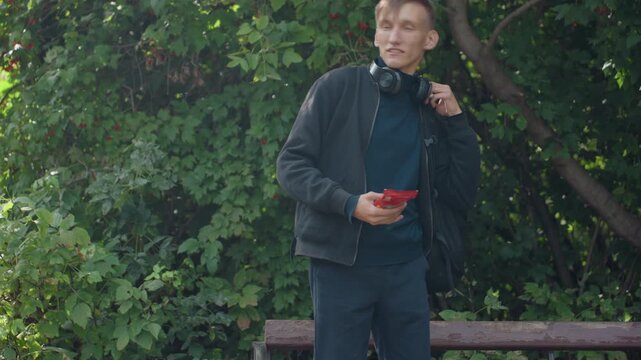 young man on bench checking phone in leafy park with backpack and headphones, sits and inspects gadget, fumbles with hands, contemplative midday mood, natural light, candid urban portrait