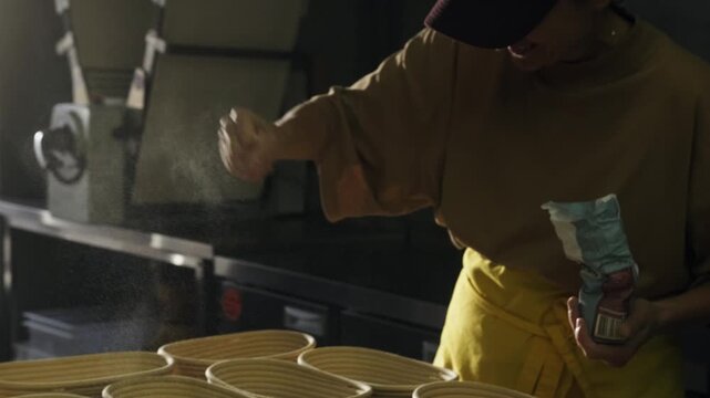 A smiling  female baker dusts bread  proofing baskets with flour in the morning sun. Flour particles float in the bakery air, artisan sourdough bread bake process.Bakery morning workflow.