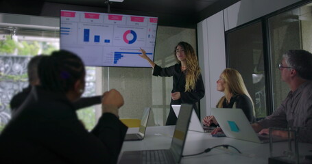 Young woman presenting data chart to colleagues while blonde businesswoman and team members listen attentively in collaborative office meeting