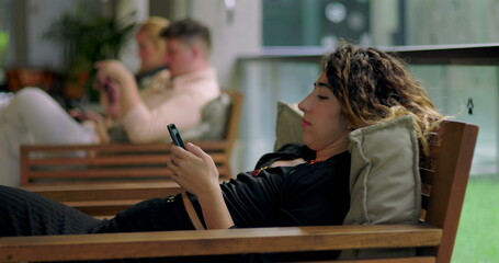 Fototapeta premium Young woman relaxing on wooden bench using smartphone indoors, with two people in background using mobile devices in a casual shared workspace