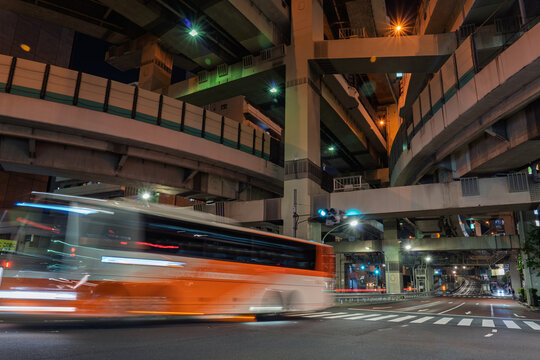 View of a blur of orange and white, a bus speeds through the streets framed by a labyrinth of concrete and steel, lit by glowing streetlights, Shuto Expressway, Tokyo, Japan.