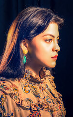 Fototapeta premium Profile of Young Latin American Woman with Long Black Hair in Dimly Lit Room Wearing Earrings and Red Lipstick