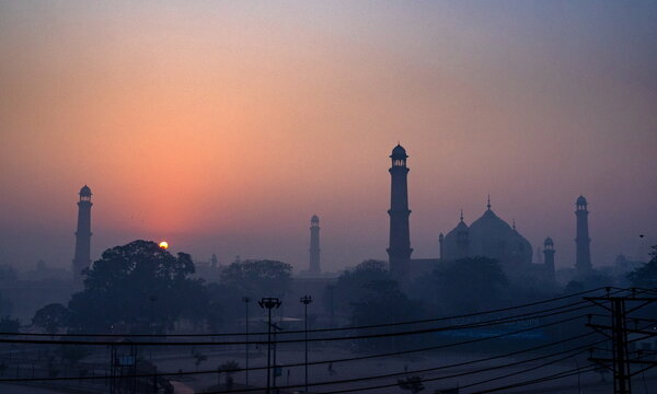 View of a hazy sunrise bathes the historic Badshahi Mosque and its minarets in soft, ethereal light, creating a serene silhouette, Lahore, Punjab, Pakistan.