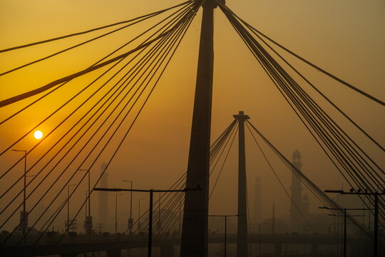 View of cable-stayed bridge silhouetted against a hazy sunrise, its cables stretching like golden threads, with distant buildings shrouded in mist, Lahore, Punjab, Pakistan.