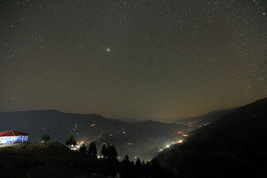 View of a starlit sky blankets the dark, mountainous landscape, with a small building illuminated on the left and distant lights twinkling below, Upper Dir, Khyber Pakhtunkhwa, Pakistan.