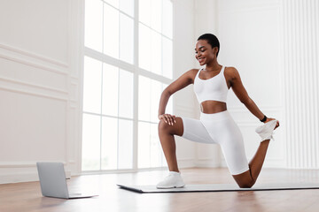 Online Training Session. Happy black fit lady in white sportswear stretching leg muscles and quads while watching online tutorial on pc, exercising in living room or fitness studio, looking at screen