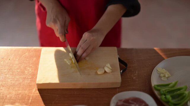 A person is cutting garlic on a wooden cutting board. The garlic is on a plate. The person is wearing a red apron