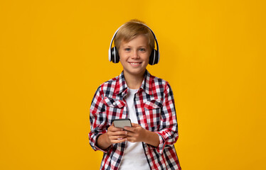 Happy european teen boy in earphones holding smartphone, watch video lesson, isolated on yellow background, copy space. Pupil listening lecture, music or audiobook. Education and technology, new app © Prostock-studio