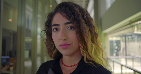 Young woman standing in modern office hallway, neutral expression, looking directly at camera with confidence, holding tablet in hands, indoor professional environment