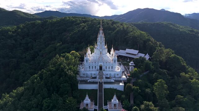 Wide drone shot of a white temple on a mountain ridge with layered hills and sky