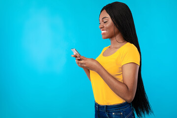 Side View Of Happy Black Woman Using Phone Texting And Browsing Internet Standing Over Blue Studio Background. New Application For Smartphone Concept, Blank Space For Advertisement Text