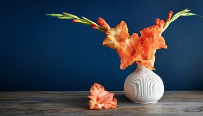 bright orange gladiolus flowers arranged in a textured white vase on a wooden table against a dark blue wall with one fallen flower beside the vase