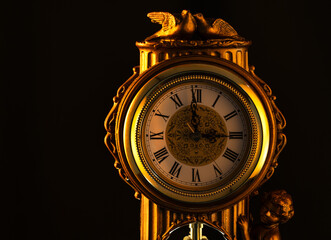 A close-up shot of an ornate golden clock with Roman numerals on its face, set against a dark background. 