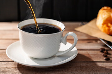 A close-up shot of someone pouring hot coffee into a white coffee cup, with bread placed on a table, in the warm morning light.