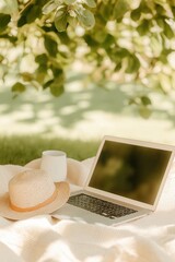 Freelancer workspace in nature. Laptop, straw hat, and mug on a cozy blanket on green grass in a park on a sunny day. Remote work concept
