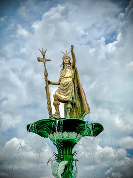 Aerial view of the golden Pachacutec statue atop a vibrant green fountain against a backdrop of swirling clouds creates a striking contrast, Cusco, Peru.