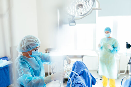 Senior female surgeon in sterile gown manages surgical cables and equipment under operating light during gynecological procedure. Experienced medical practice, teamwork in modern operating room. - Powered by Adobe