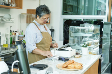 Asian senior female barista in apron organizing receipts at cafe counter smiling gently managing tasks preparing orders enjoying independent post-retirement life running small family coffee business.