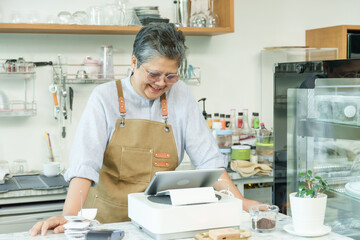 Asian senior female cafe owner smiling leaning on counter using tablet enjoying working confidently running independent small coffee shop demonstrating post-retirement happiness in business.