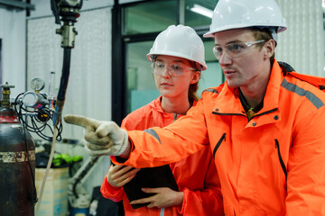 Caucasian male engineer wearing safety gear instructing asian female trainee inside smart factory using robotic welding arm in automation training session for workforce skill development