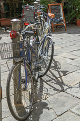 Obraz premium Row of bicycles locked to post on a sunny Italian street with stone pavement and cafe sign. Urban cycling, eco-friendly transport, lifestyle, travel, and European city environment.