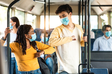 New Normal Greeting. Diverse multiethnic people doing elbow tap bumping standing together at in public transport, wearing single-use surgical masks. Asian guy and black woman avoid coronavirus spread