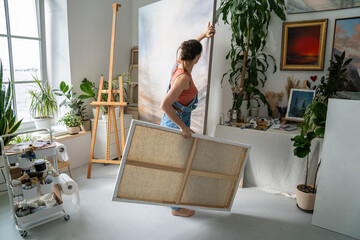 Focused woman painter working on painting in studio, placing canvas on easel adjusting artworks,...