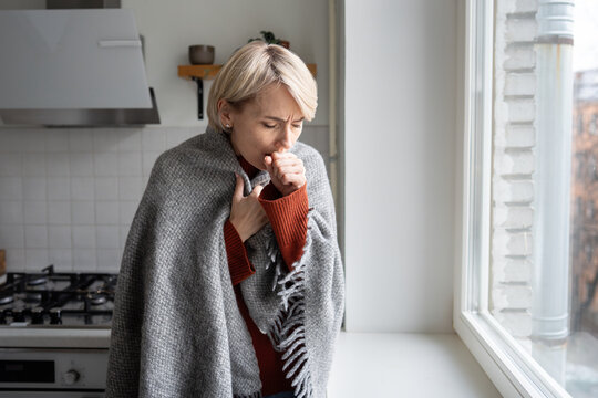 Unhealthy woman wrapped in knitted plaid with sick expression stands near window, signs of cold and flu, heavy coughing and sore throat symptoms caused by weather changes and seasonal infection.