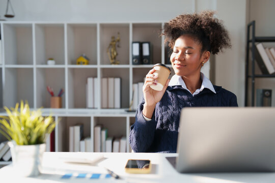 African American woman having a coffee break during work, smiling with closed eyes, appreciating the warm beverage at her office desk with a laptop and shelves in the background