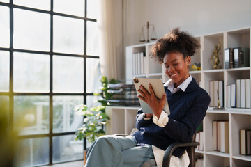 Young African American woman smiling while working on a digital tablet, managing business in a professional law firm office with bookshelves and a window © Tj