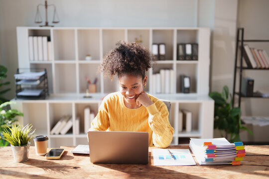 Young African American woman in a yellow sweater smiling as she types on a laptop at a bright wooden desk, focused and productive in a modern home office setting