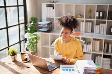 Young Black woman at desk happily reviewing paperwork and financial charts, organizing stacked...