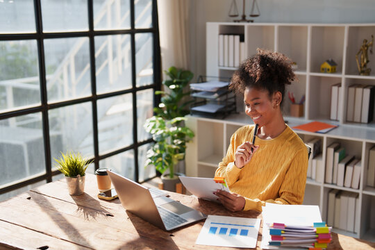 Young happy African American woman reviewing financial documents and planning strategy at her desk, with a laptop and organized files in a modern office environment
