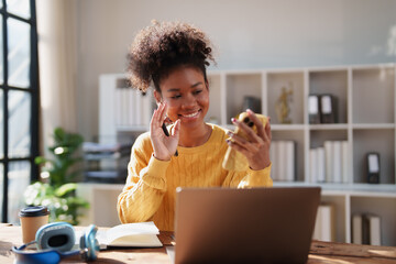 Young black woman waving and smiling into a smartphone screen, engaging in a video call or online meeting while working remotely from a modern home office with laptop and headphones on the desk